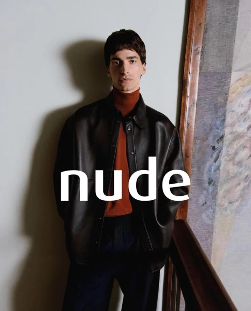 Portrait of a brunette man in a dark leather jacket and orange turtleneck, standing against a neutral background with a painting.