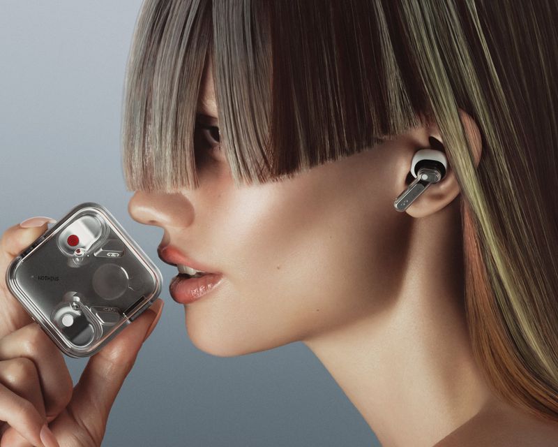 A fair-skinned woman with blunt bangs is holding a Nothing ear (2) case while wearing a white and silver Nothing ear (2) earbud.