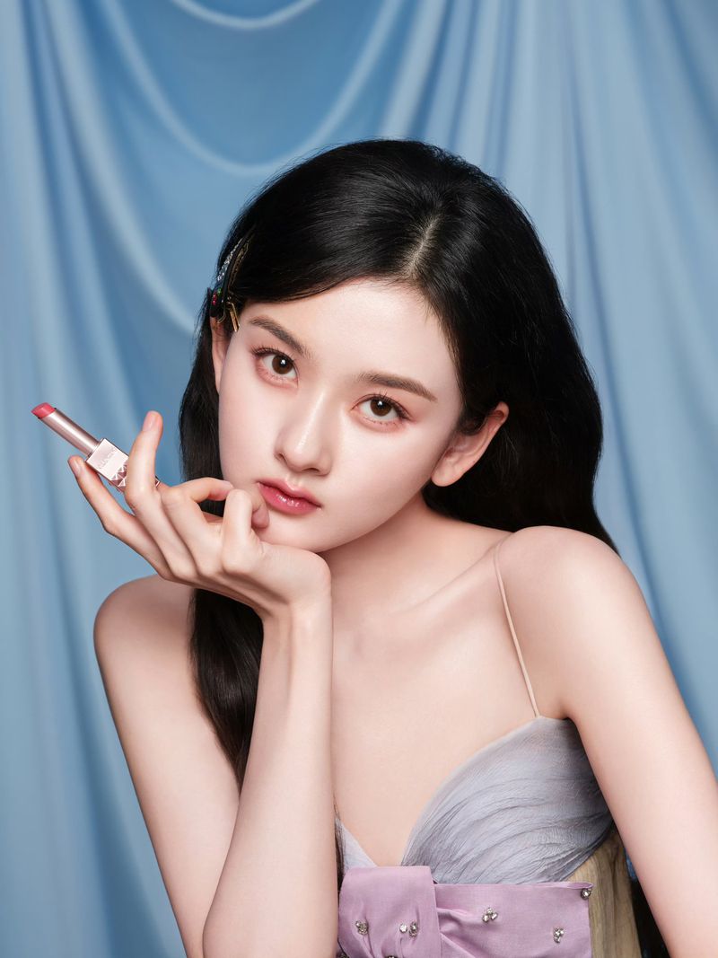Close-up of a woman with dark hair holding lipstick, wearing a formal gown in front of a light blue draped fabric backdrop.