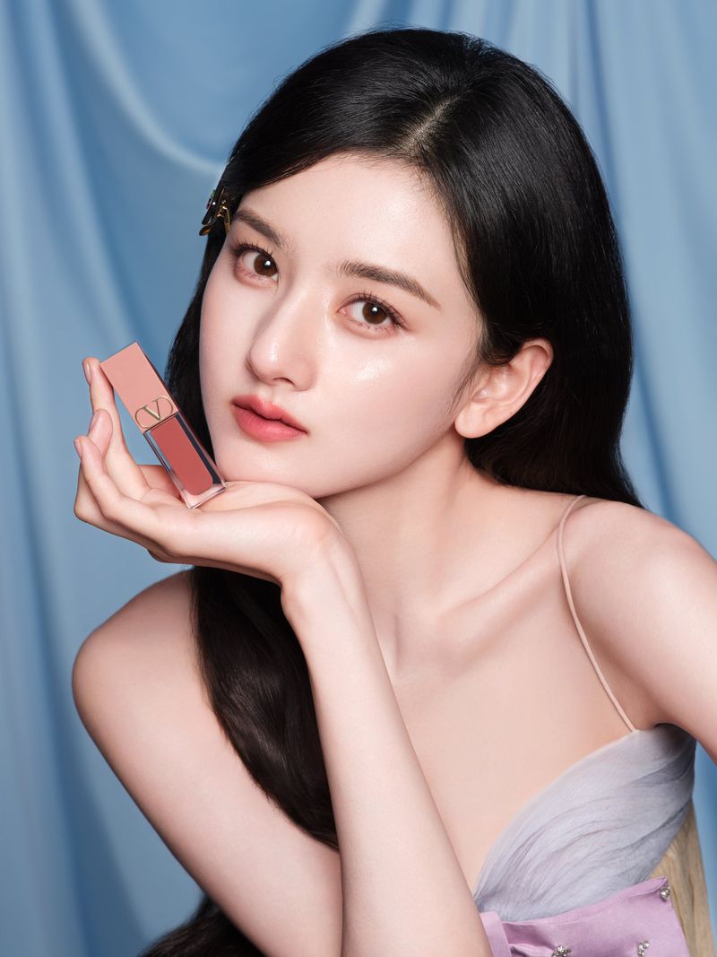 Portrait of a young Asian woman with long black hair, holding a pink lipstick tube in front of a draped light blue background.
