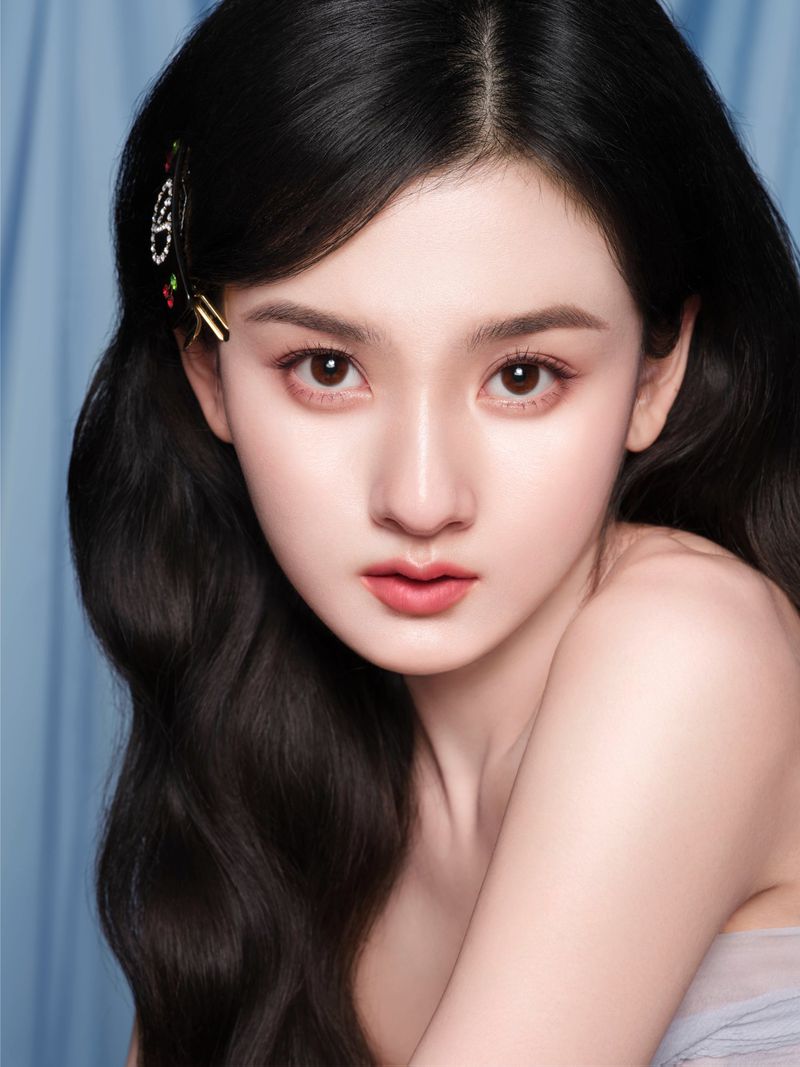 Close-up portrait of an Asian woman with long, dark, wavy hair, fair skin, and a hair clip, set against a soft blue background.