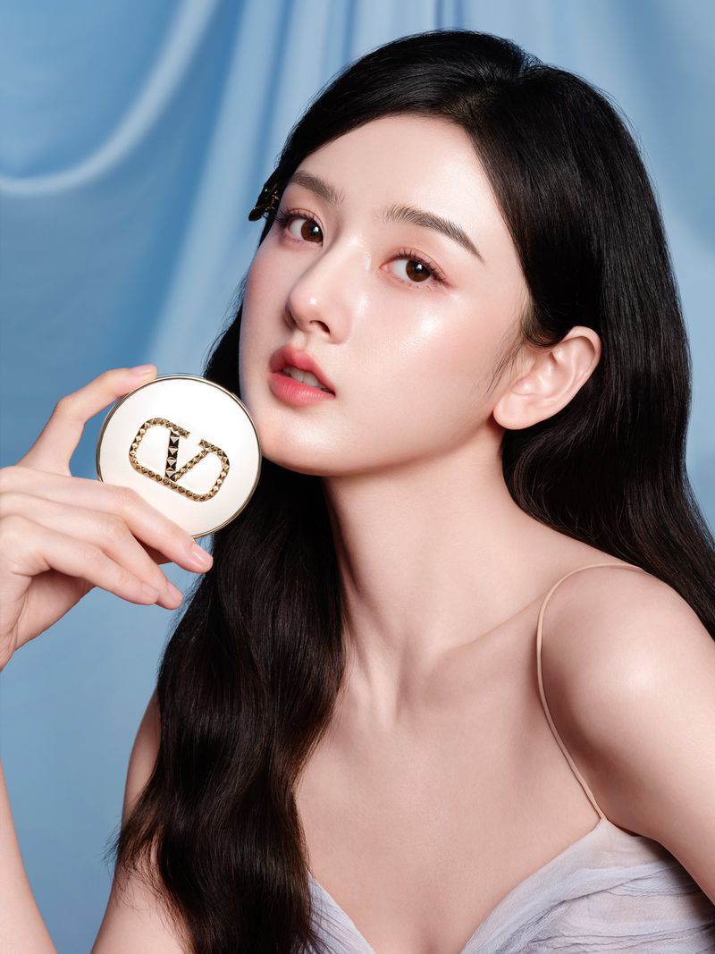 Close-up studio shot of a woman with dark hair holding a makeup compact in front of a blue fabric background, beauty concept.
