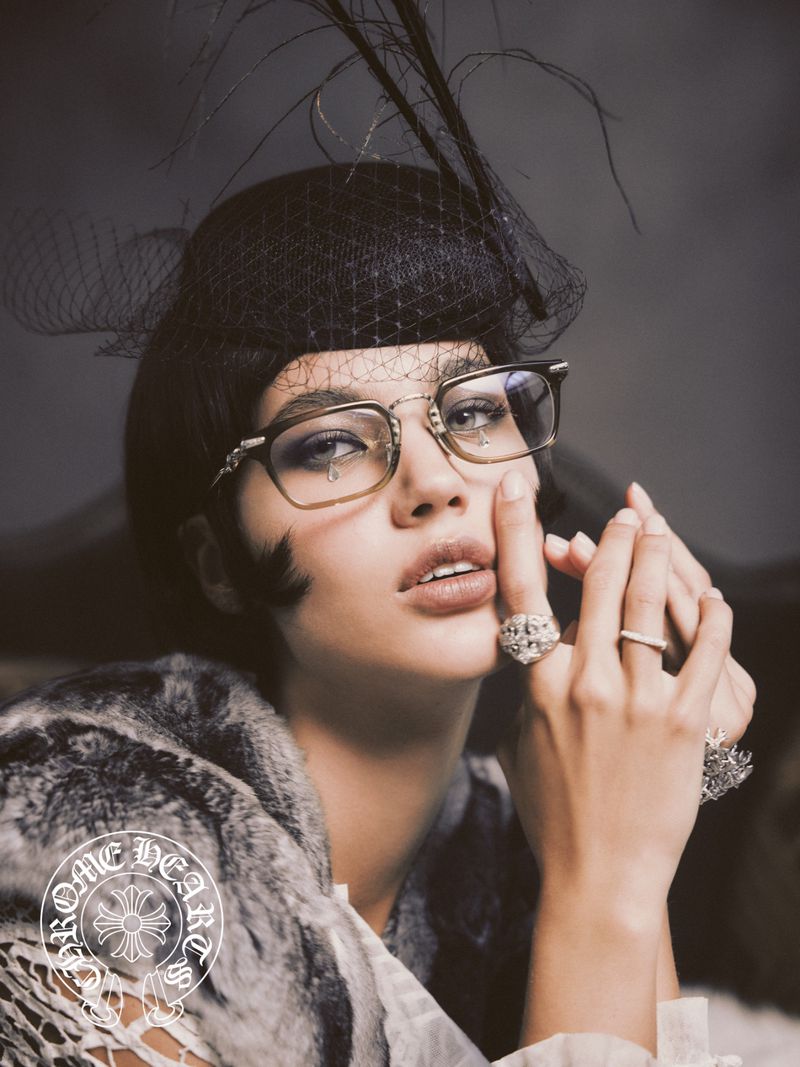 Portrait of a woman wearing glasses and a black feathered hat. She has rings on her fingers and is wearing a fur stole.
