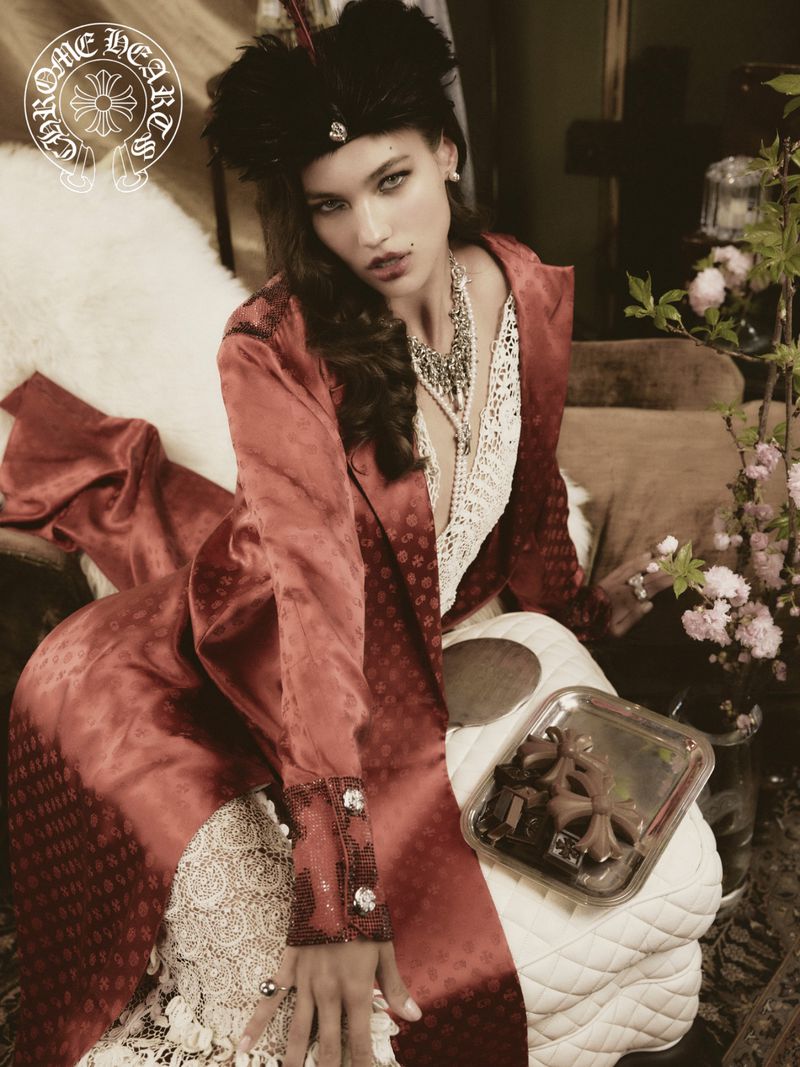 Elegant woman with feathered hat and ornate jewelry, wearing a patterned coat and lace dress in a styled indoor fashion portrait.