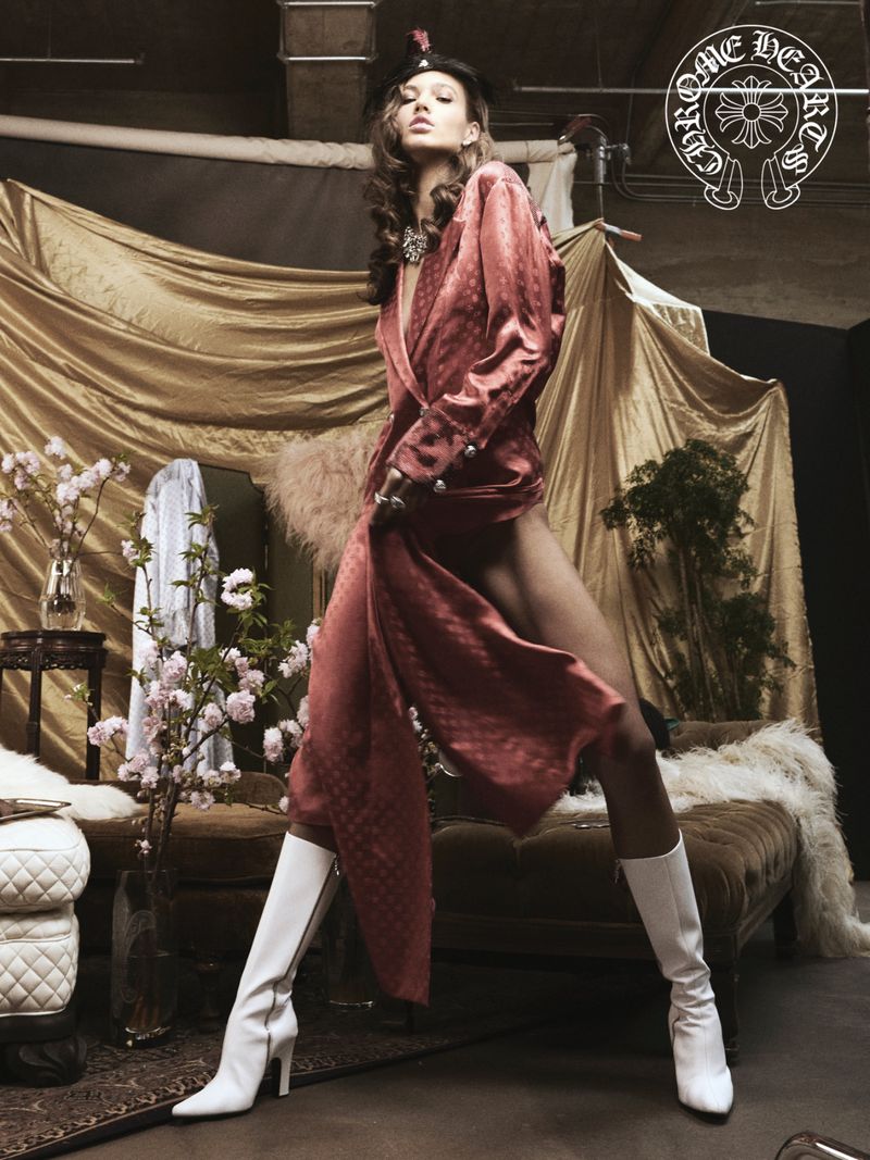 A fashionably dressed woman poses in white knee-high boots and a robe in a studio, standing in front of a gold draped backdrop.