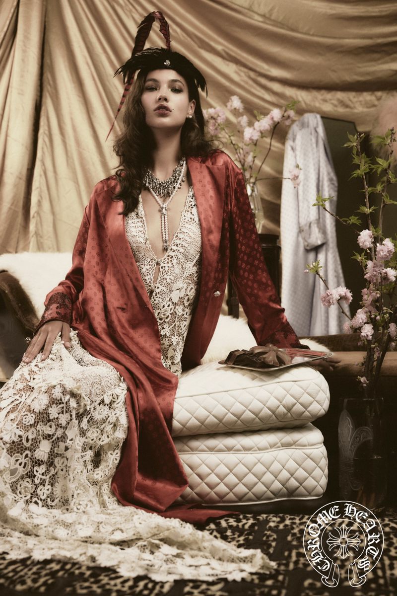 A woman in a red robe and feathered hat sits in an indoor fashion shot with visible jewelry, cushions, and a decorative floral arrangement.