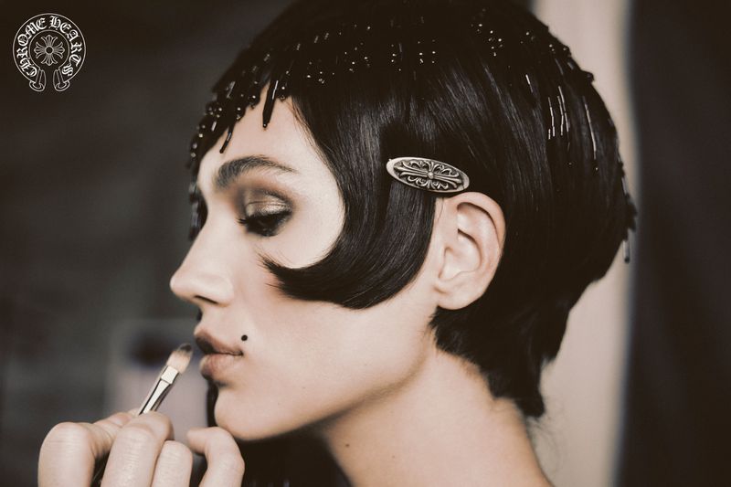 Close-up of a woman with bobbed hair and a beaded headpiece getting makeup applied with a brush in a muted, dark environment.