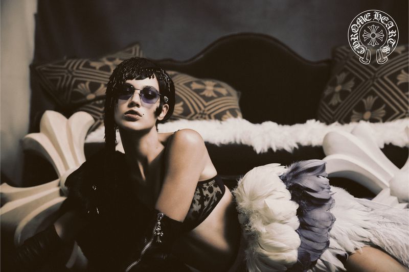 Adult woman with sunglasses lies on a couch wearing a bra and black gloves. The couch is ornately decorated with patterned pillows.