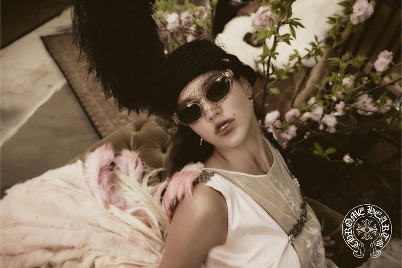 Woman in vintage-style sunglasses, hat with feather, and dress poses on a velvet couch amid flowers, evoking a nostalgic fashion aesthetic.