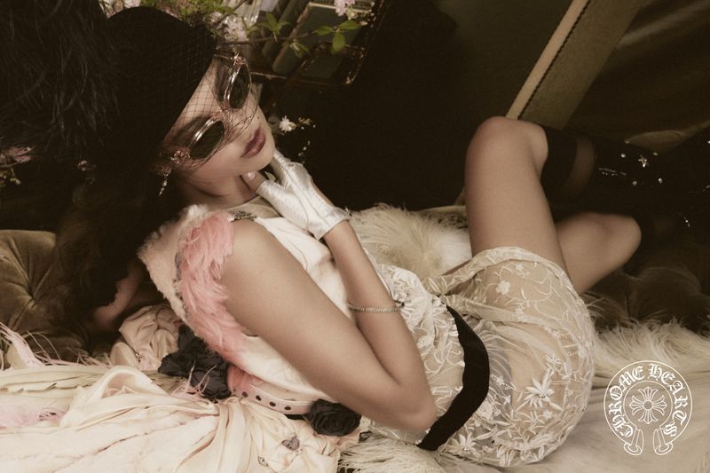 Woman in vintage-inspired fashion reclines among soft fabrics wearing a feathered hat, round sunglasses, gloves, and sheer floral dress.
