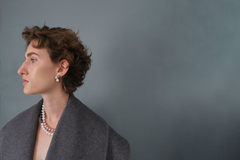 Elegant woman in gray coat, side profile, wearing pearl necklace and earrings against a muted blue background in a studio portrait.