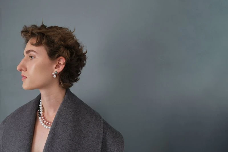 Elegant woman in gray coat, side profile, wearing pearl necklace and earrings against a muted blue background in a studio portrait.