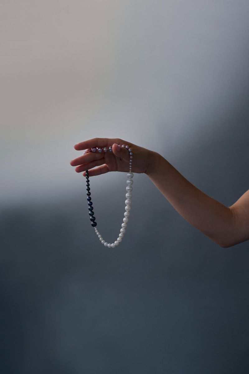 A hand holding a dangling necklace crafted with white pearls and a twisted black accent against a muted grey studio backdrop.