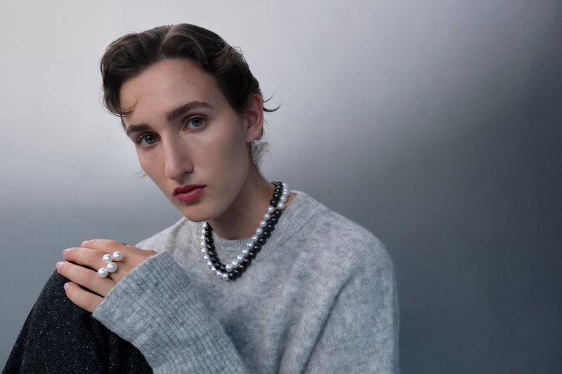 Person with pearl jewelry: close-up of a young adult wearing a pearl necklace and ring with a gray sweater against a gradient gray backdrop.