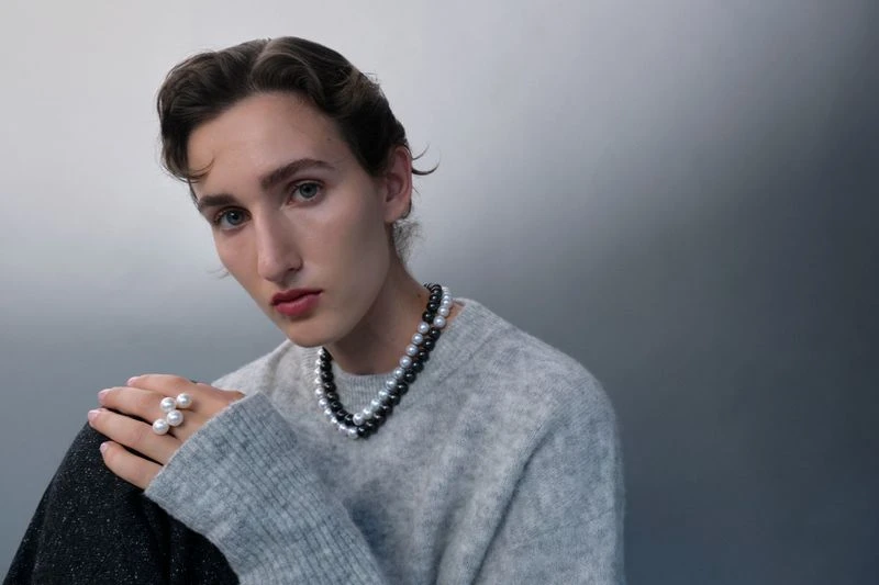 Person with pearl jewelry: close-up of a young adult wearing a pearl necklace and ring with a gray sweater against a gradient gray backdrop.