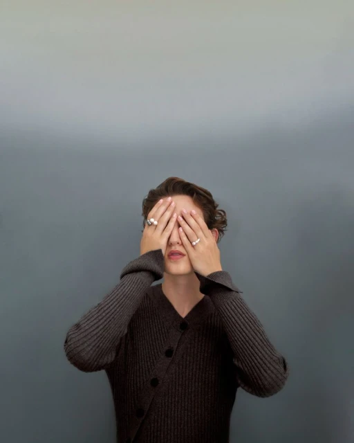 Young adult with hands covering face, wearing a dark sweater with a button closure, against a smooth gradient backdrop, medium shot.