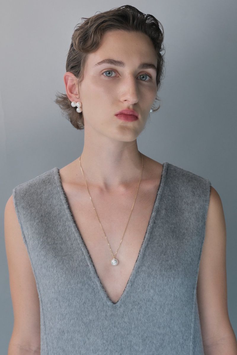 Close-up of a fair-skinned person with pearl jewelry, wearing a sleeveless grey top, against a light grey background.