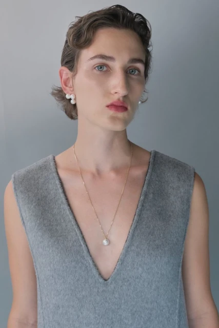 Close-up of a fair-skinned person with pearl jewelry, wearing a sleeveless grey top, against a light grey background.