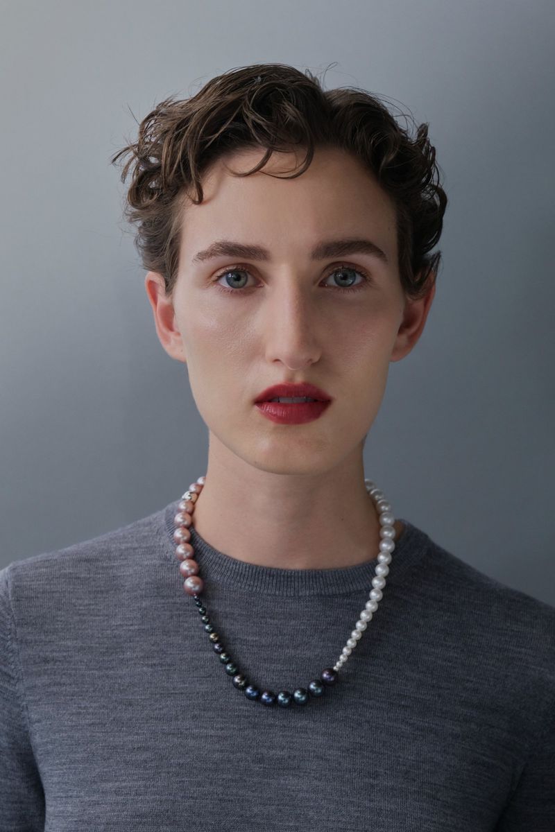 Close-up portrait of an adult woman with short curly hair, fair skin, and a pearl necklace, set against a neutral grey backdrop.