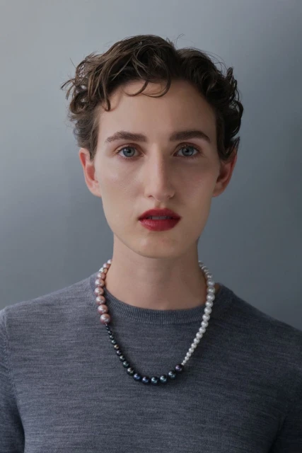 Close-up portrait of an adult woman with short curly hair, fair skin, and a pearl necklace, set against a neutral grey backdrop.