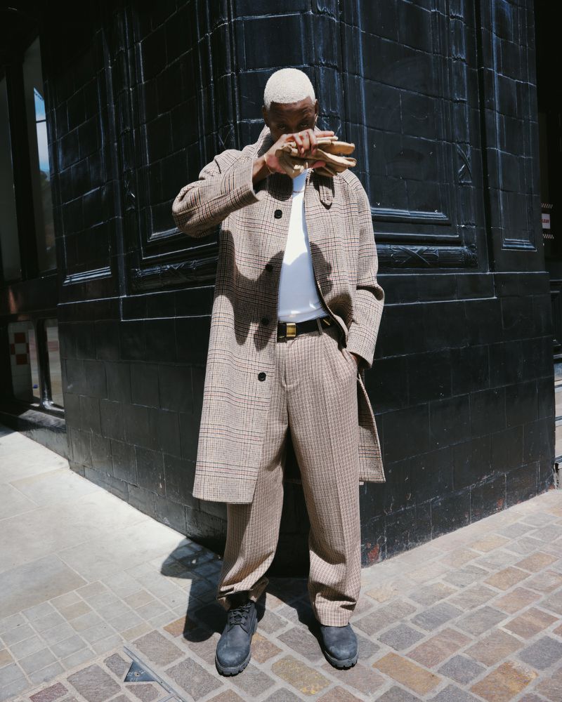 Fashionable adult male with bleached hair, wearing a plaid overcoat, pants, and boots, standing on a city sidewalk in daylight.
