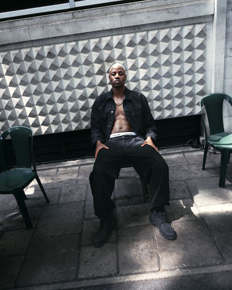 Man with shaved head sits on chair outdoors wearing black jacket, pants, and boots in front of textured concrete wall.