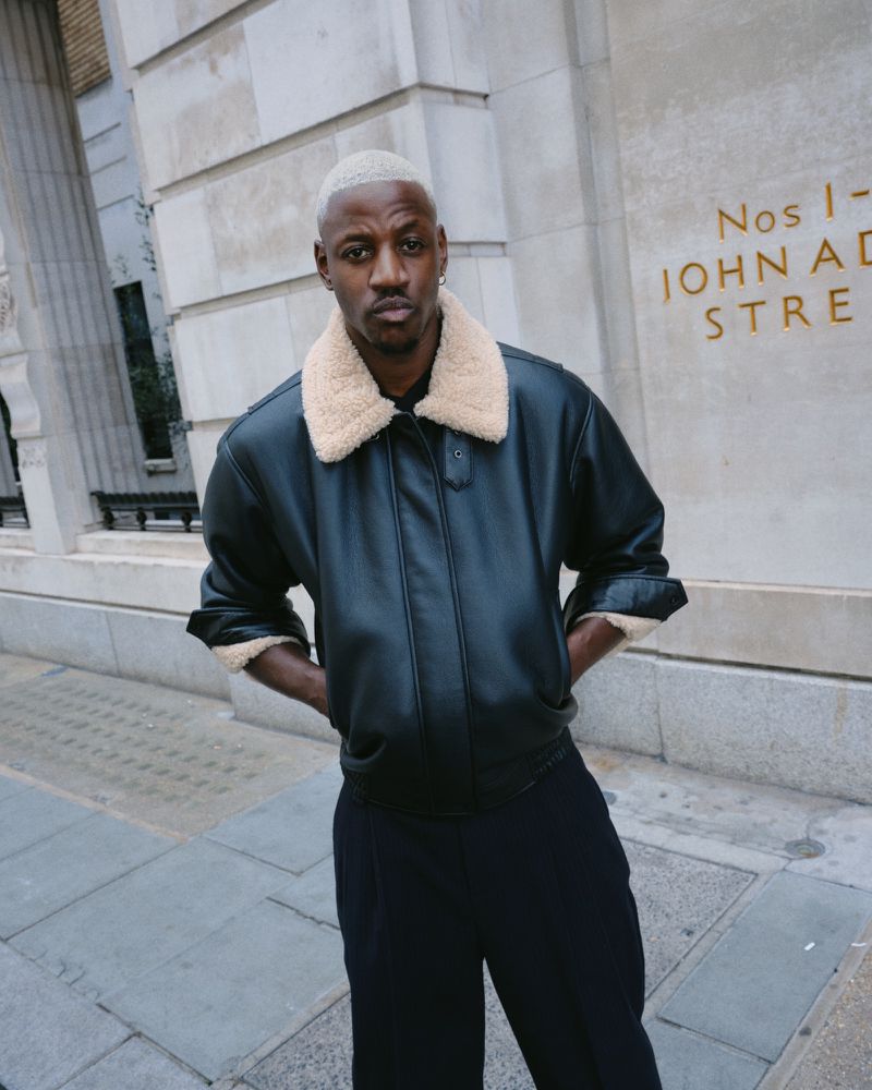 Adult male with light blonde hair poses in a black leather jacket with a shearling collar and dark pants against a light stone wall.