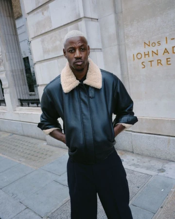 Adult male with light blonde hair poses in a black leather jacket with a shearling collar and dark pants against a light stone wall.