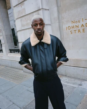 Black man with bleached hair wearing a black leather bomber jacket with a cream fleece collar on an urban street.