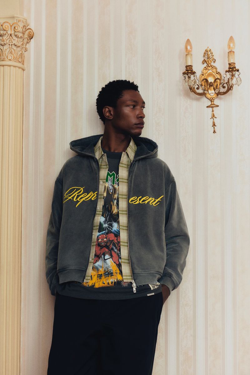 Man in gray Represent brand zip-up hoodie, layered shirt, and black pants poses indoors by decorative wall and ornate sconce lighting.