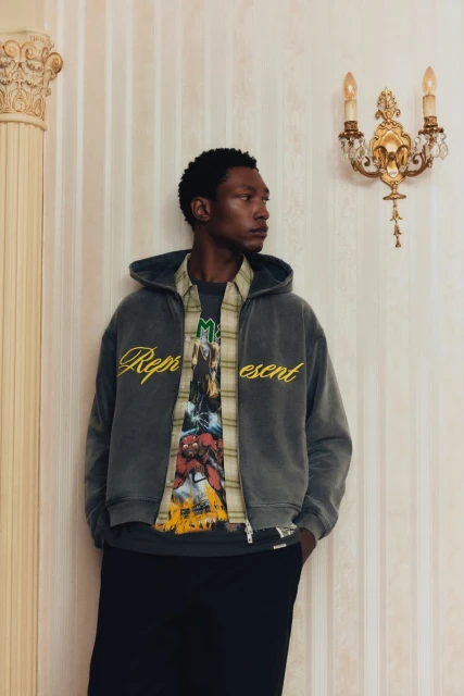 Man in gray Represent brand zip-up hoodie, layered shirt, and black pants poses indoors by decorative wall and ornate sconce lighting.