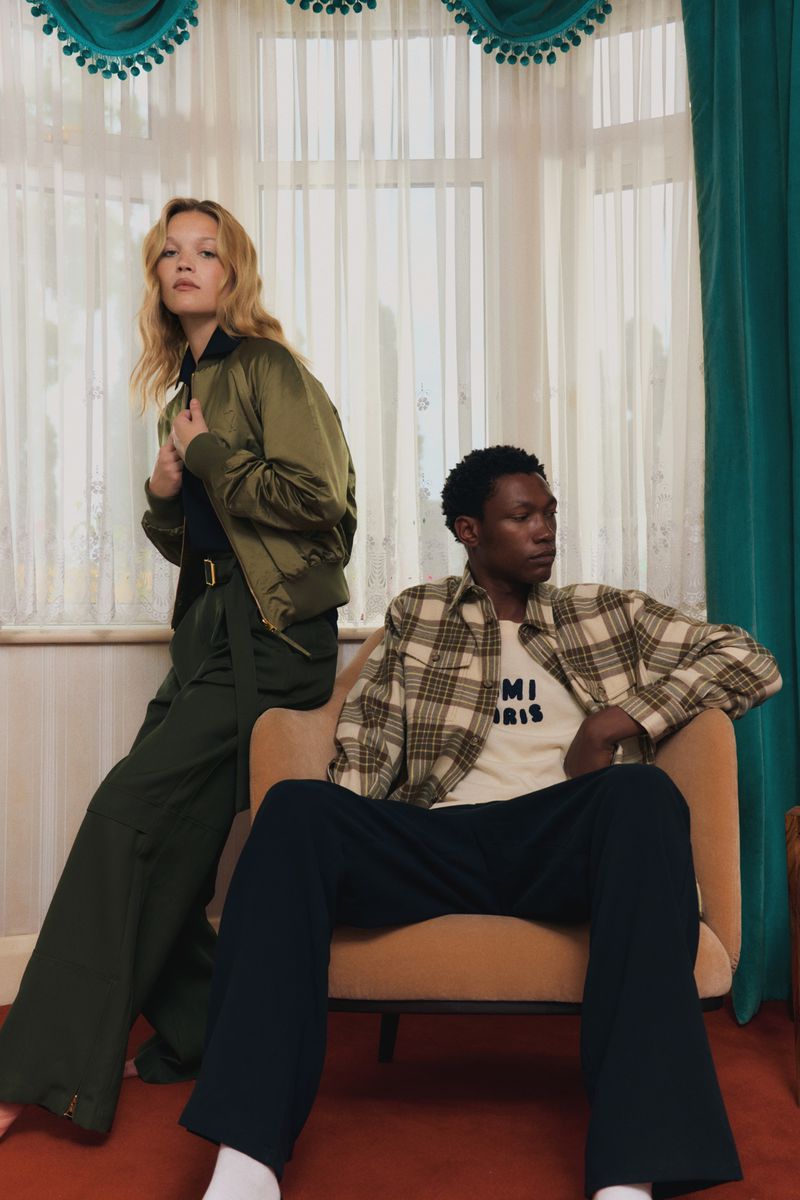 A blond woman and a Black man in fashion clothing are in an indoor setting; she wears a bomber jacket, and he sits in a chair.