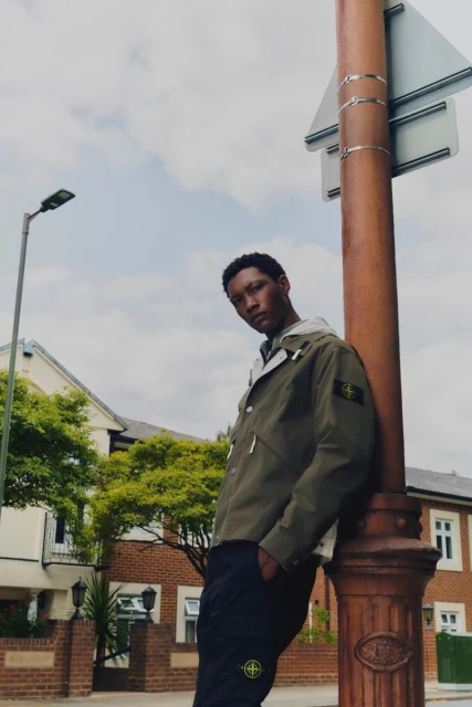 Young man in olive green jacket leans against pole outdoors, with residential buildings in background on a cloudy day, fashion shoot.