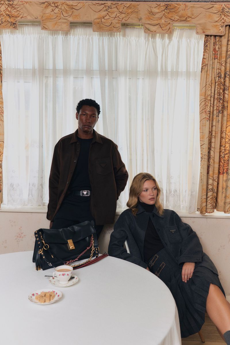 Two fashion models pose indoors near a table with tea and a handbag. The setting features curtains and neutral wall colors.