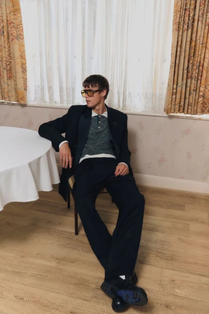 Man in a suit and yellow glasses sits at a table in an indoor setting with a patterned wall, bathed in soft, diffused light.