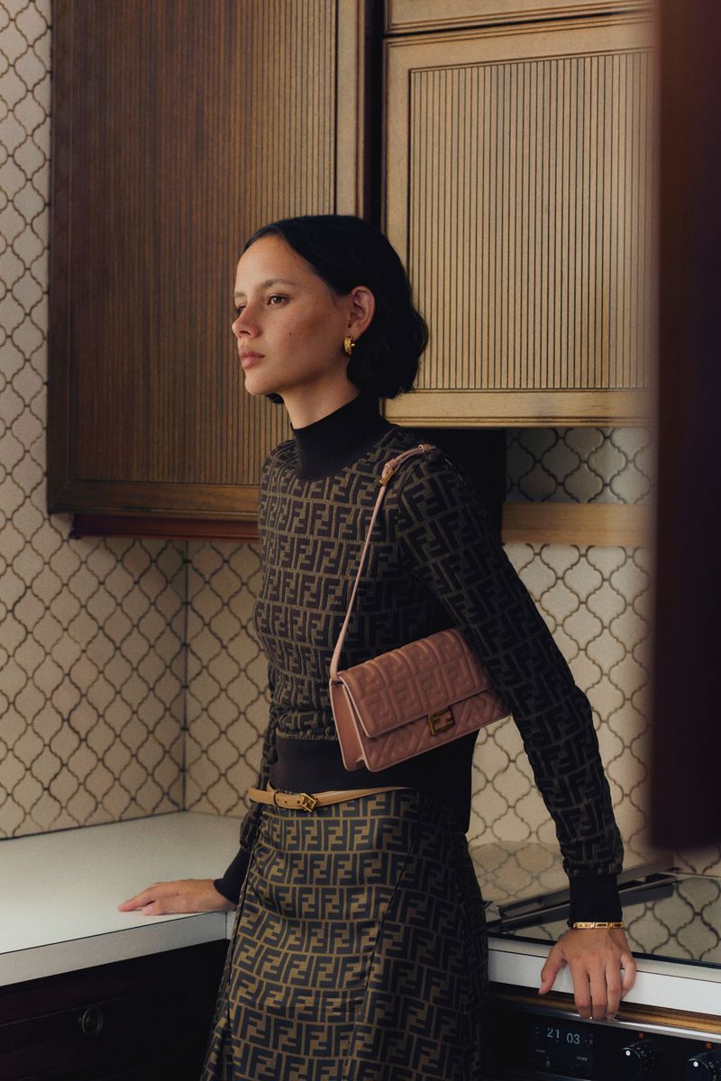 Model standing in a domestic kitchen wearing monogram print turtleneck and skirt, carrying a pink shoulder bag.