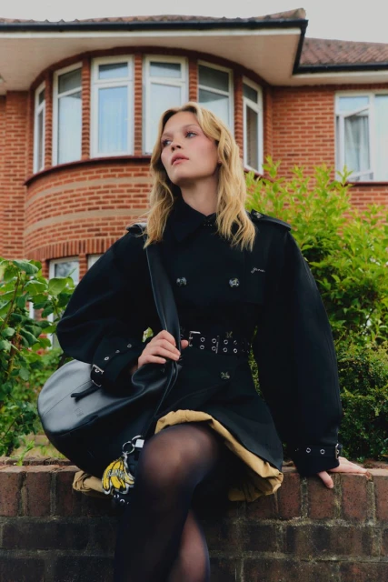 Adult female sitting on a brick wall outside a brick house, wearing a black coat, skirt, tights and carrying a black bag. 