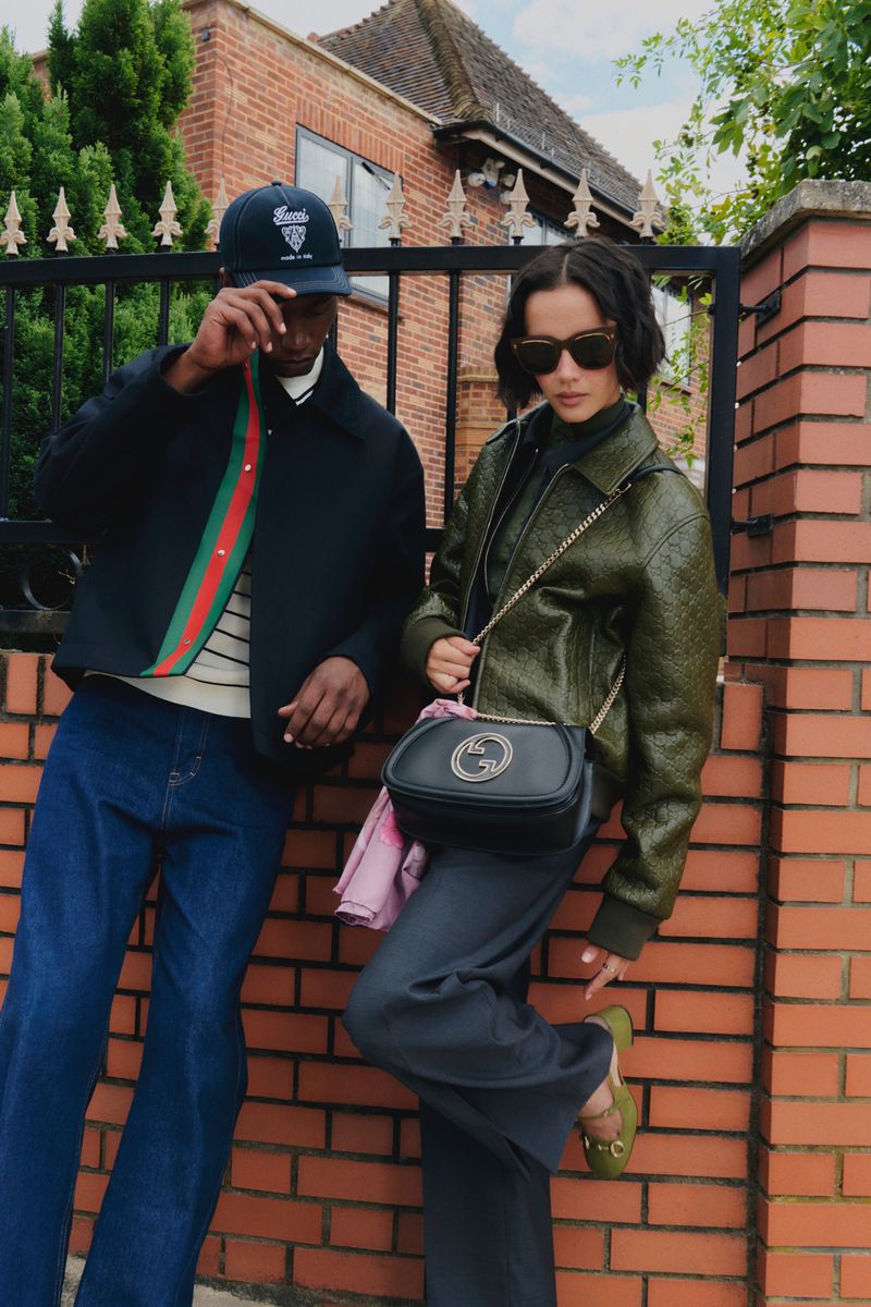An adult couple, dressed in fashionable jackets and trousers, pose against a brick wall outside of a brick house with a metal fence.