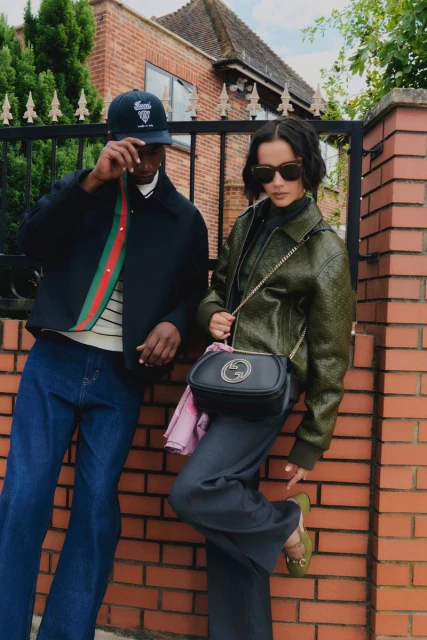 An adult couple, dressed in fashionable jackets and trousers, pose against a brick wall outside of a brick house with a metal fence.