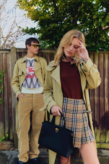 Two fashion models in trench coats and plaid skirts and pants pose outdoors with a fence, greenery, and sky in the background.