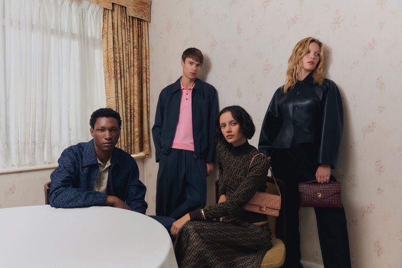Four fashion models pose indoors against a floral wallpaper backdrop wearing stylish clothing, with a table in the foreground.