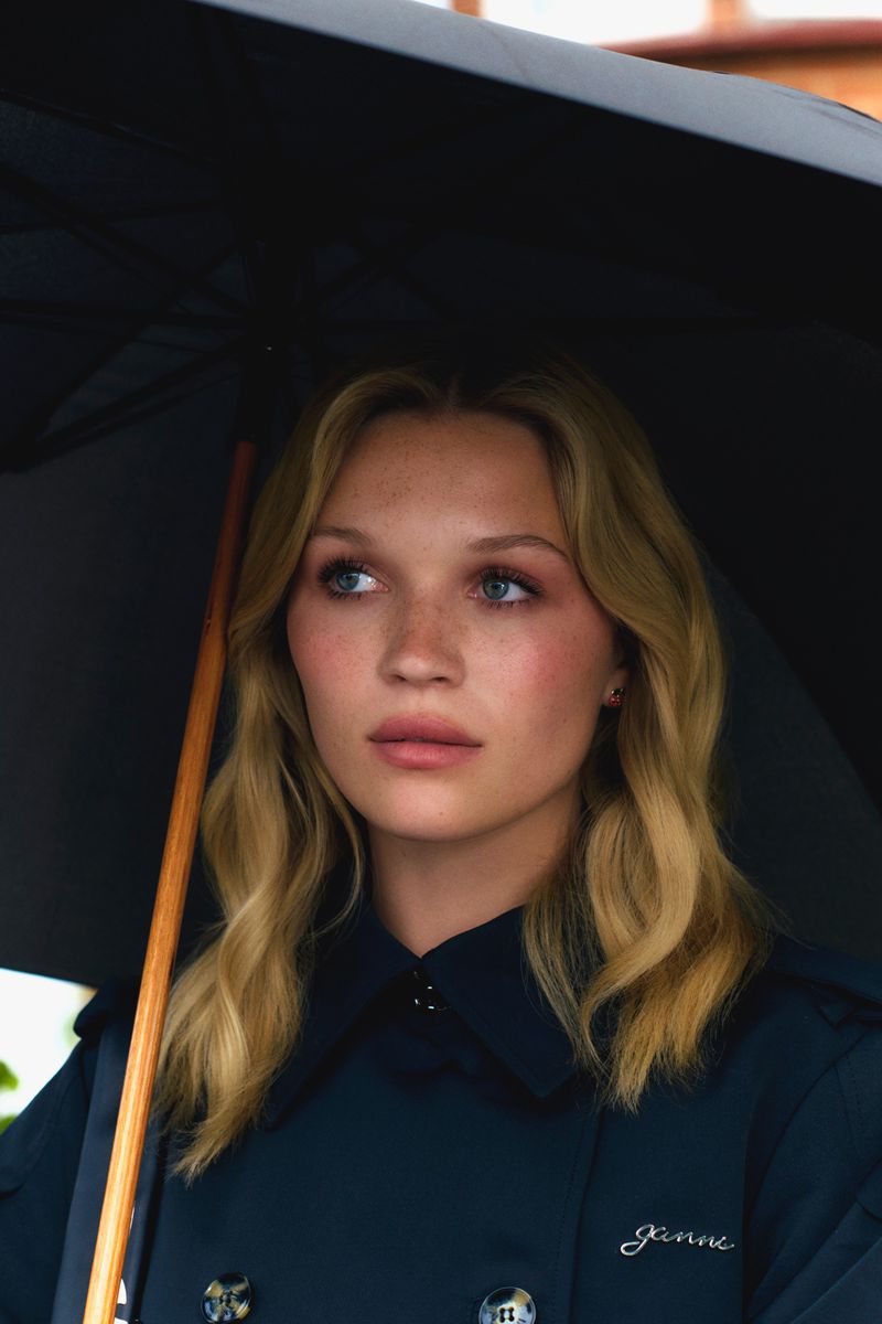 Blonde woman with fair skin, wearing a blue coat and standing under a black umbrella, looks to the left in a close-up shot.