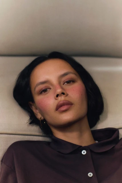 Portrait of a young woman with short dark hair wearing a brown collared shirt, showcasing a clean beauty look and soft studio lighting.