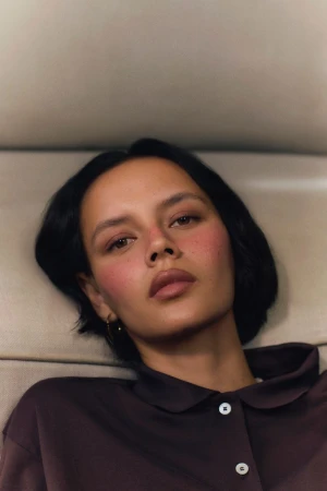 Portrait of a young woman with short dark hair wearing a brown collared shirt, showcasing a clean beauty look and soft studio lighting.