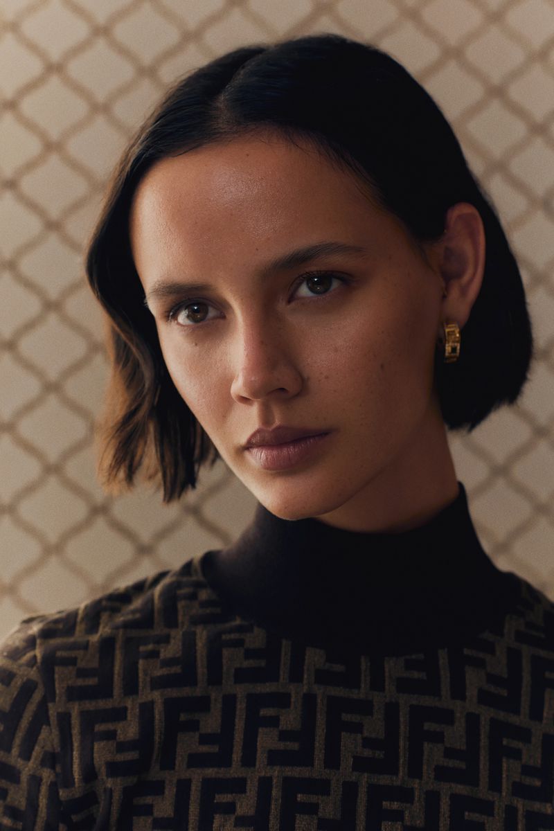 Close-up portrait of a young adult female with dark hair, gold earrings, and a patterned brown and black turtleneck sweater against a patterned background.