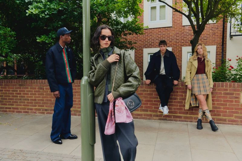 Four people in stylish fall outfits pose in front of a brick building. The woman in the middle wears a quilted green jacket and sunglasses.
