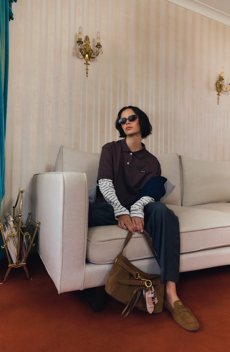 Woman in brown polo shirt, striped long-sleeve shirt, and sunglasses sits on a white couch with a brown suede handbag.