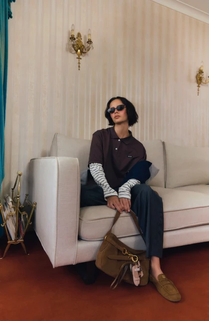 Woman in brown polo shirt, striped long-sleeve shirt, and sunglasses sits on a white couch with a brown suede handbag.
