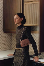 Model standing in a domestic kitchen wearing monogram print turtleneck and skirt, carrying a pink shoulder bag.