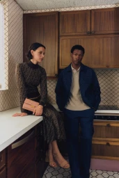 A woman in a patterned brown knit set and a man in a navy jacket posing inside a retro wood-paneled kitchen.